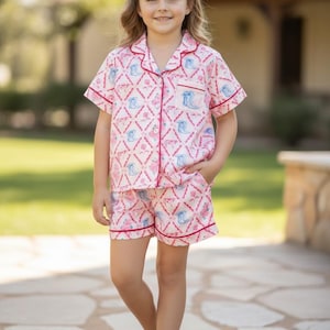 K&ouml;nnte beinhalten: Rosa-wei&szlig;es Pyjama-Set mit einem Muster aus blauen Cowboy-Stiefeln und rosa Rosen. Das kurz&auml;rmelige Oberteil und die Shorts haben einen roten Besatz. Das Kind steht auf einem Steinweg.