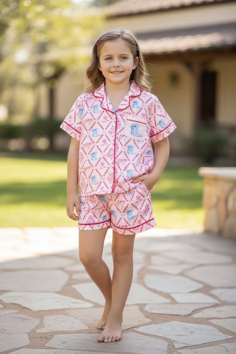 K&ouml;nnte beinhalten: Rosa-wei&szlig;es Pyjama-Set mit einem Muster aus blauen Cowboy-Stiefeln und rosa Rosen. Das kurz&auml;rmelige Oberteil und die Shorts haben einen roten Besatz. Das Kind steht auf einem Steinweg.