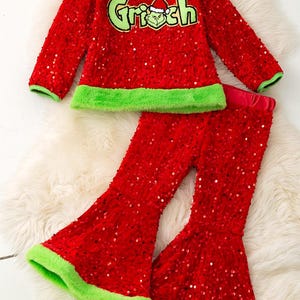 May include: A red sequined Grinch-themed outfit, including a long-sleeved top and flared pants. The top features the word "Grinch" in green with a Grinch face graphic. Green trim accents the neckline, cuffs, and pants hem.
