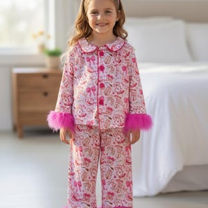 以下が含まれることがあります： ピンク色のパジャマセット。リボンとギフトの繰り返し模様。長袖トップスとパンツは、袖口と足首に明るいピンク色のフェイクファーのトリムが付いています。お揃いのヘアバンドがコーディネートを完成させます。
