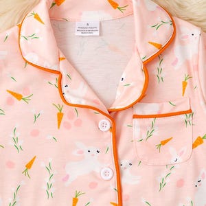 Peut inclure: Haut de pyjama rose clair avec un imprim&eacute; sur le th&egrave;me de P&acirc;ques. Le motif comprend des lapins blancs, des carottes oranges et des brins verts. Le haut a une bordure orange, une poche poitrine et des boutons blancs.