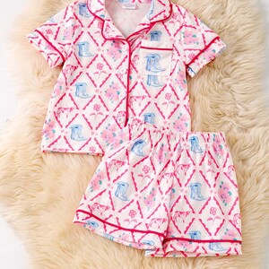K&ouml;nnte beinhalten: Ein zweiteiliger Schlafanzug mit einem sich wiederholenden Muster aus rosa Blumen, blauen Cowboy-Stiefeln und rosa Rauten auf wei&szlig;em Grund. Das Set besteht aus einem kurz&auml;rmeligen Hemd mit Kn&ouml;pfen und passenden Shorts, beide mit roter Paspel versehen.