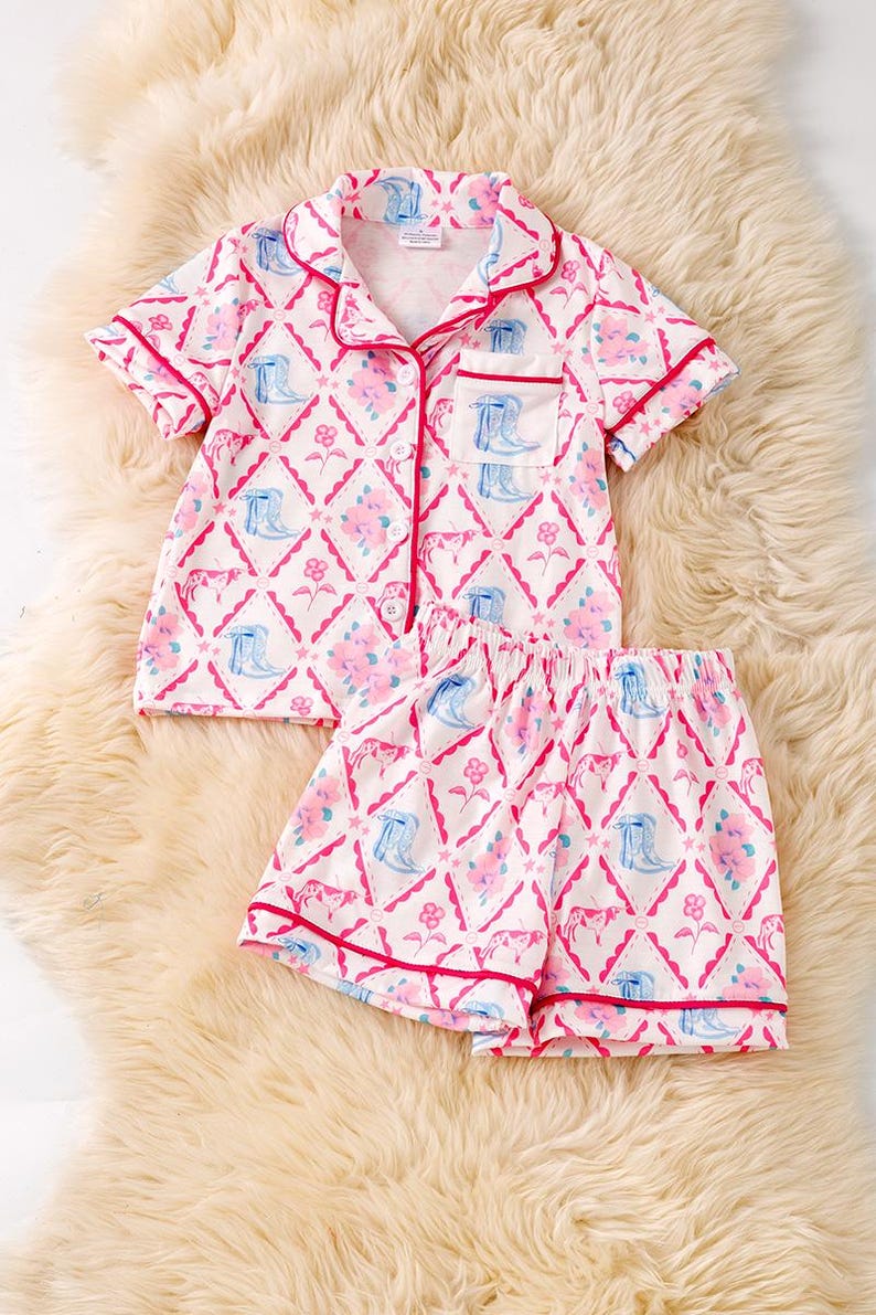 K&ouml;nnte beinhalten: Ein zweiteiliger Schlafanzug mit einem sich wiederholenden Muster aus rosa Blumen, blauen Cowboy-Stiefeln und rosa Rauten auf wei&szlig;em Grund. Das Set besteht aus einem kurz&auml;rmeligen Hemd mit Kn&ouml;pfen und passenden Shorts, beide mit roter Paspel versehen.