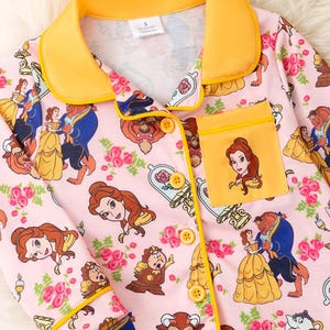 May include: Pink pajama set featuring a repeating pattern of Belle, the Beast, and other characters from Beauty and the Beast, with rose and floral accents. The set has a yellow collar, pocket, and trim, with yellow buttons.