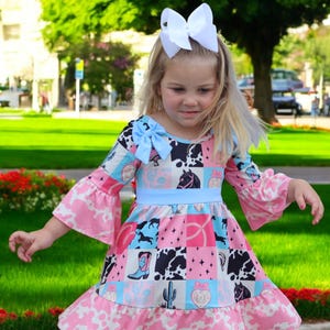 以下が含まれることがあります： ピンク、青、黒のパッチワークドレスを着た若い女の子。カウガールをテーマにしたドレスで、ベルスリーブ、水色のリボン、髪には白いリボンが付いています。背景は緑の芝生と木がある公園です。