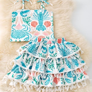 Puede incluir: Conjunto de dos piezas con estampado de vida marina. La parte superior blanca tiene tirantes anudados y la falda tiene tres capas con volantes y borlas blancas. El estampado incluye langostas, conchas y coral en verde azulado, coral y rosa.