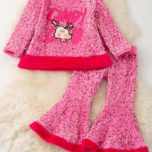 May include: A pink sequined two-piece outfit featuring a long-sleeved top and flared pants. The top has a red trim and the words "Cindy Lou" with a cartoon character. The pants also have red trim.