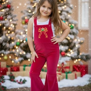 May include: A young girl wearing a red velvet jumpsuit with a bow design on the bib. She is wearing a white tank top and white sneakers. The background features Christmas trees and wrapped gifts.