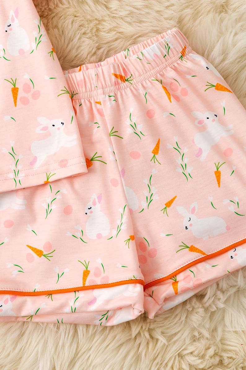 Peut inclure: Ensemble de pyjama rose avec un motif de lapins blancs, de carottes oranges et de brins verts. Le short et le haut ont une bordure orange. Le tissu est doux et l&eacute;ger, parfait pour le printemps ou P&acirc;ques.