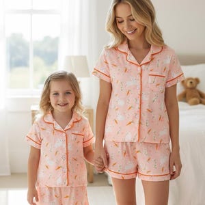 Peut inclure: Ensembles de pyjamas roses assortis avec un motif de lapin et de carottes. Les ensembles comprennent une chemise boutonn&eacute;e &agrave; manches courtes et un short, bord&eacute;s d'un passepoil orange. Une femme et un enfant portent les pyjamas.