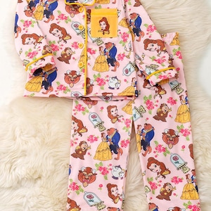 May include: Pink pajama set featuring a repeating pattern of characters and imagery from Beauty and the Beast. The set includes a button-down top with a yellow collar and pocket, and matching pants. Yellow trim accents the edges.