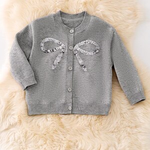Könnte beinhalten: Graue Strickjacke mit Pailletten-Schleifenmuster. Der Pullover hat einen runden Ausschnitt, lange Ärmel und eine Knopfleiste. Die Schleife besteht aus silbernen Pailletten. Die Strickjacke wird auf einer flauschigen, cremefarbenen Oberfläche präsentiert.