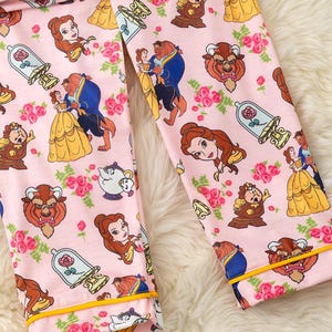 May include: Pink pajama pants featuring a repeating pattern of characters and elements from the animated film, Beauty and the Beast. The design includes Belle, the Beast, roses, and other iconic imagery. The pants have yellow trim.