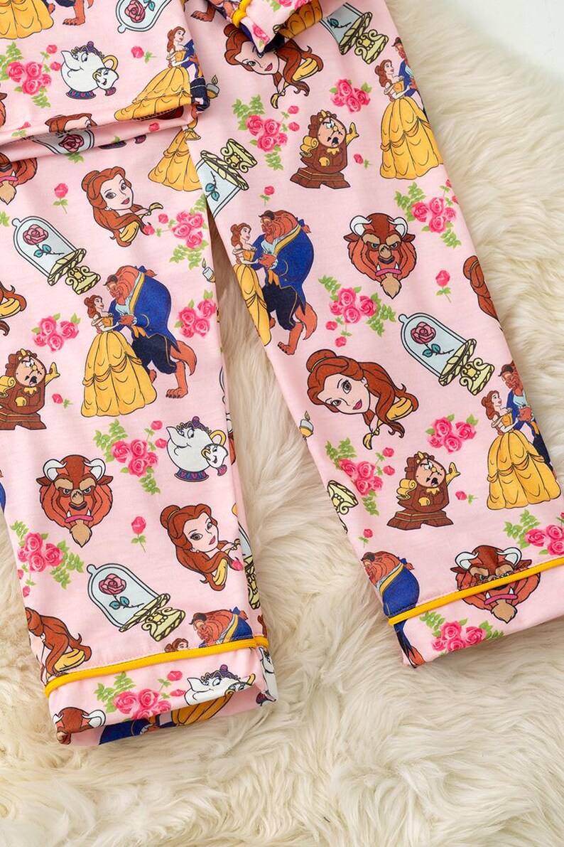 May include: Pink pajama pants featuring a repeating pattern of characters and elements from the animated film, Beauty and the Beast. The design includes Belle, the Beast, roses, and other iconic imagery. The pants have yellow trim.