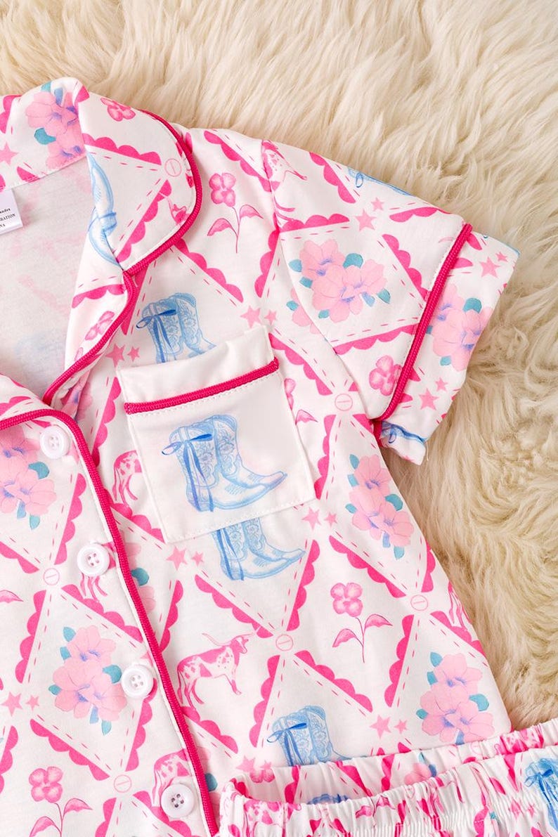 K&ouml;nnte beinhalten: Wei&szlig;es Pyjama-Set mit einem sich wiederholenden Muster aus rosa Blumen, blauen Cowboy-Stiefeln und rosa Sternen. Die Paspel ist hellrosa, und die Tasche zeigt eine Cowboy-Stiefel-Illustration. Das Set hat ein Oberteil mit Kn&ouml;pfen und Shorts.