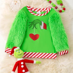 May include: A green Christmas-themed sweater with a Grinch design, featuring a hand holding a red heart. The sweater has fluffy green sleeves and red and white striped trim. A Grinch plush toy and a beaded necklace are also in the image.