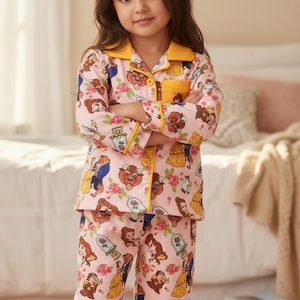 May include: Pink pajama set featuring Beauty and the Beast characters. The long-sleeved top has a yellow collar and trim, with a matching pocket. The pants and top are covered in a repeating pattern of the characters and roses.