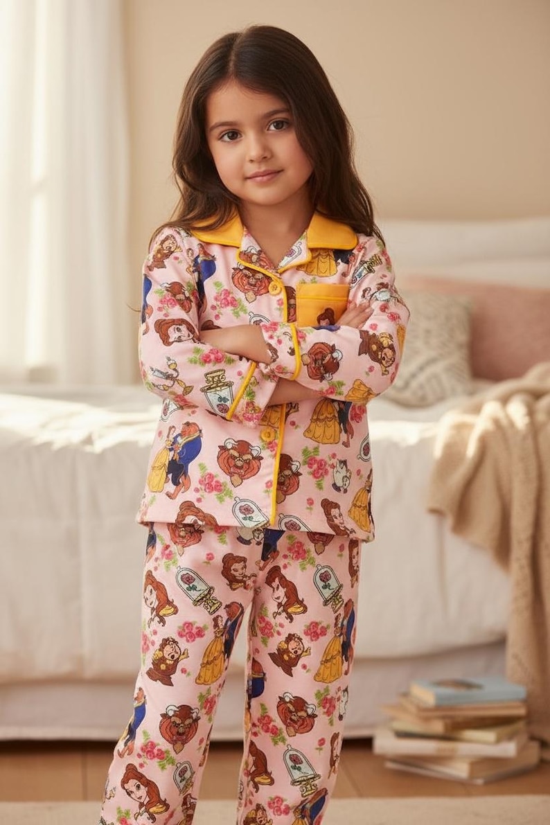 May include: Pink pajama set featuring Beauty and the Beast characters. The long-sleeved top has a yellow collar and trim, with a matching pocket. The pants and top are covered in a repeating pattern of the characters and roses.