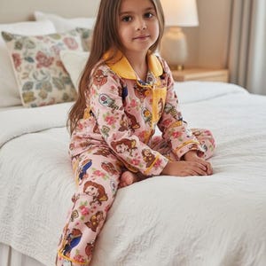 May include: Pink pajama set featuring a floral and character print. The long-sleeved top and pants have yellow trim. The pajamas are worn by a person sitting on a white bedspread.