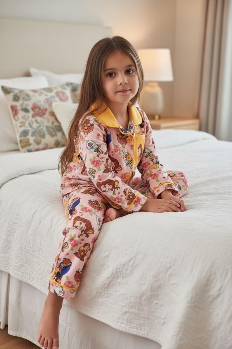 May include: Pink pajama set featuring a floral and character print. The long-sleeved top and pants have yellow trim. The pajamas are worn by a person sitting on a white bedspread.
