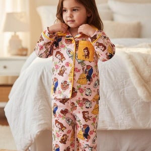 May include: Pink pajama set featuring a repeating pattern of characters from Beauty and the Beast, including Belle, the Beast, and Mrs. Potts. The set has yellow trim and a pocket with Belle's image. The pajamas are worn by a young child.