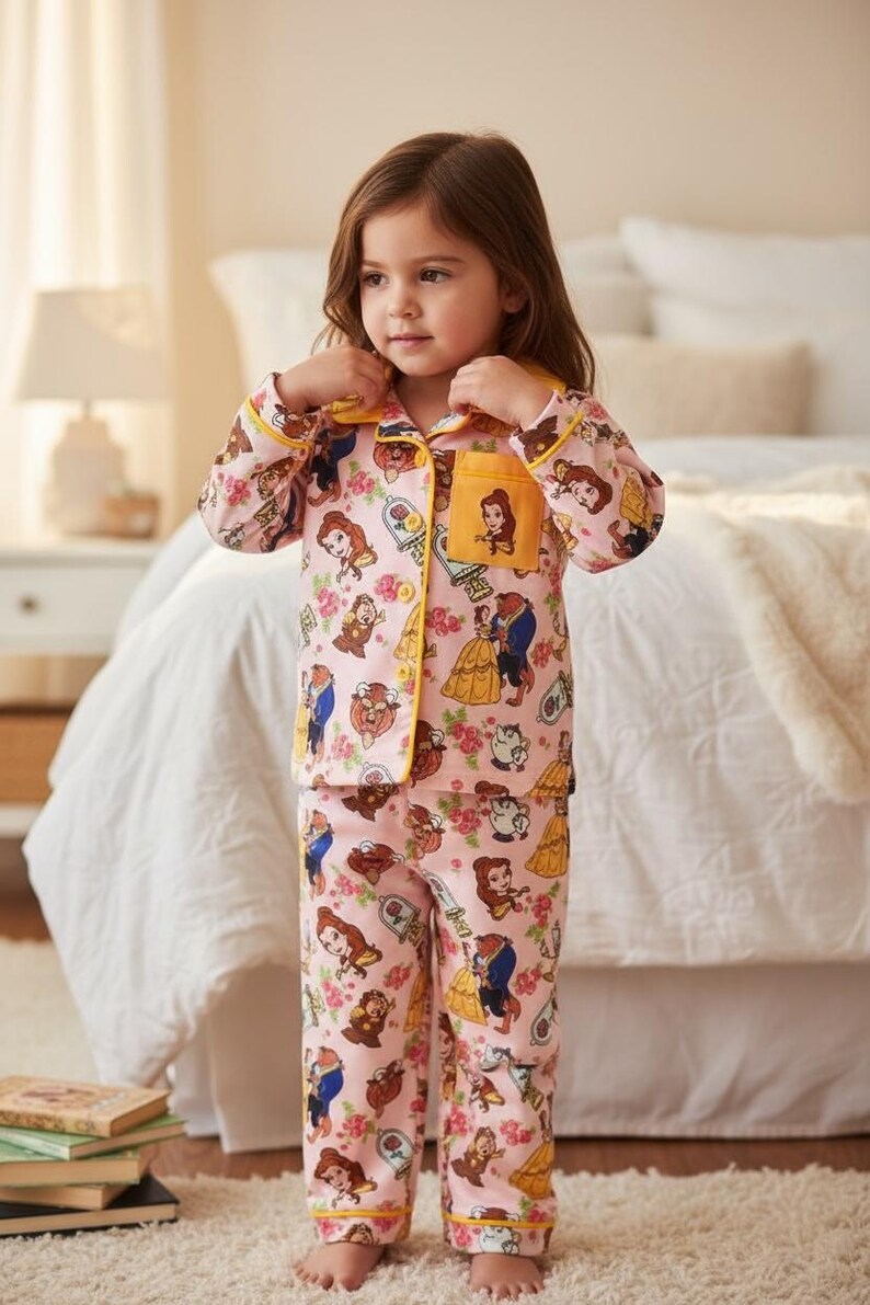 May include: Pink pajama set featuring a repeating pattern of characters from Beauty and the Beast, including Belle, the Beast, and Mrs. Potts. The set has yellow trim and a pocket with Belle's image. The pajamas are worn by a young child.