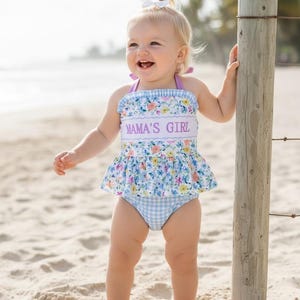 May include: A baby girl's swimsuit with a floral and gingham pattern. The top features the text "MAMA'S GIRL" in purple. The swimsuit has a ruffled skirt and is trimmed with lavender. The baby is standing on a sandy beach.