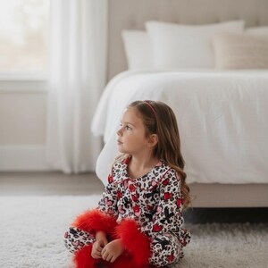 Pu&ograve; includere: Una persona giovane indossa un pigiama con stampa a cuore e leopardo. Il pigiama ha colletto e polsini rossi. La persona tiene in mano un oggetto rosso e soffice, forse una sciarpa o una boa, mentre &egrave; seduta su un tappeto bianco.