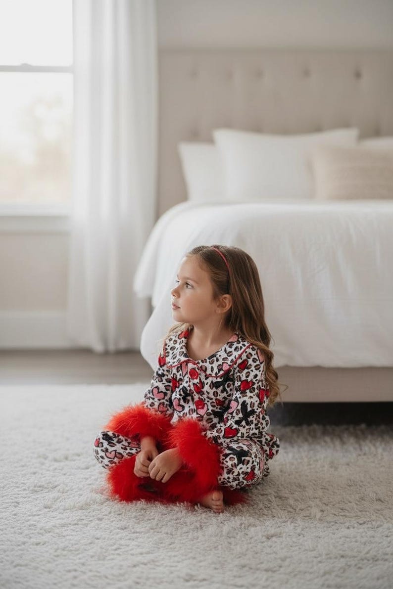 Pu&ograve; includere: Una persona giovane indossa un pigiama con stampa a cuore e leopardo. Il pigiama ha colletto e polsini rossi. La persona tiene in mano un oggetto rosso e soffice, forse una sciarpa o una boa, mentre &egrave; seduta su un tappeto bianco.