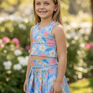 May include: A young girl in a light blue floral print crop top and pleated skirt. The outfit features pink and white flowers and green leaves. The summer outfit is ideal for warm weather and outdoor activities.