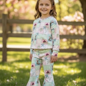 May include: A young person wearing a matching pastel-colored outfit. The long-sleeved top and flared pants feature a patchwork design with cowboy boots, hats, and bows. The outfit is in shades of pink, green, and white.