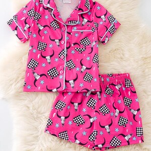 Pode incluir: Conjunto de pijama rosa com uma parte superior de manga curta abotoada e cal&ccedil;&otilde;es. A parte superior e os cal&ccedil;&otilde;es t&ecirc;m um padr&atilde;o xadrez preto e branco com um design de caveira e chifres, estrelas e raios.