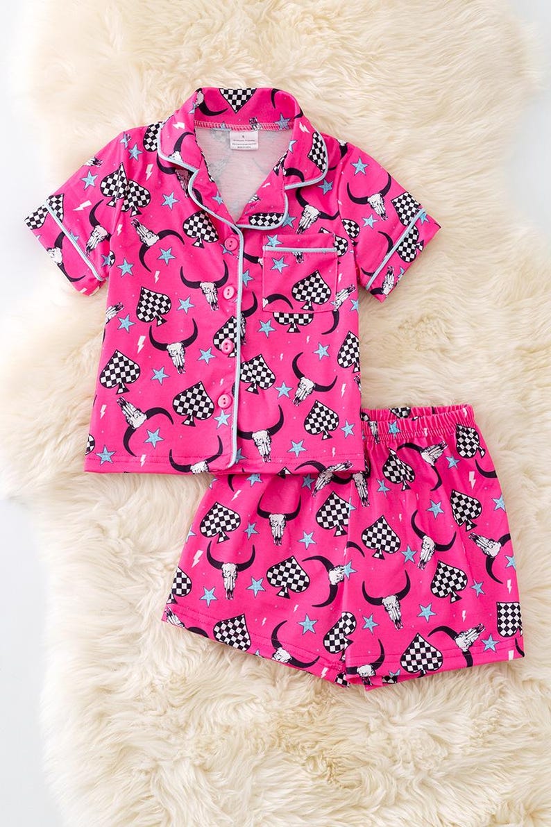 Pode incluir: Conjunto de pijama rosa com uma parte superior de manga curta abotoada e cal&ccedil;&otilde;es. A parte superior e os cal&ccedil;&otilde;es t&ecirc;m um padr&atilde;o xadrez preto e branco com um design de caveira e chifres, estrelas e raios.