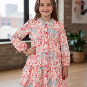 May include: A button-down dress with a pink and white pattern featuring cacti, hearts, and suns. The dress has long sleeves, a ruffled hem, and light blue accents on the collar and buttons. The girl is wearing a bracelet.