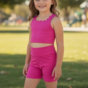 May include: A young girl wearing a bright pink two-piece outfit. The top is a crop top with a scalloped neckline, and the shorts are high-waisted. She also wears a matching pink headband. The background is a park.