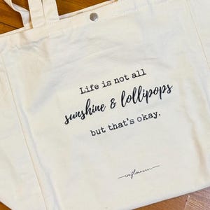 Può includere: Una borsa tote in tela bianca con testo nero che recita "Life is not all sunshine & lollipops but that's okay." Il testo è in un font scritto a mano ed è centrato sulla borsa. La borsa ha due lunghi lacci e una chiusura a bottone.
