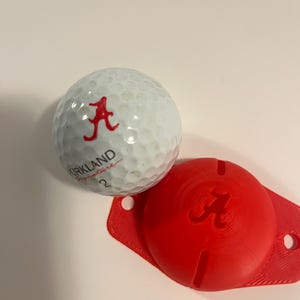 May include: A white golf ball with the text "KIRKLAND Signature 2" and a red "A" logo, next to a red golf ball marker with the same "A" logo. The golf ball and marker are on a white surface.