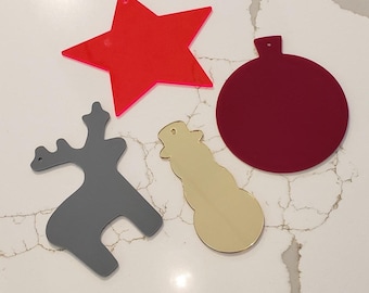 May include: Four Christmas ornaments made of acrylic. A red star, a burgundy circle, a gray reindeer, and a gold snowman.