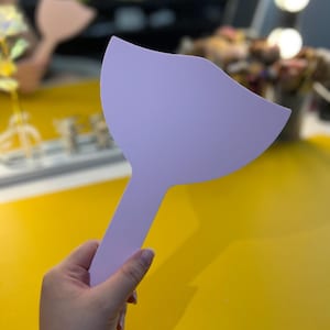 Puede incluir: Una espátula de maquillaje de plástico de color lila claro con una hoja redondeada y curvada y un mango largo.