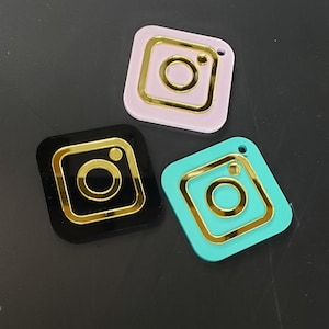 May include: Three square-shaped Instagram logo charms with gold outlines. The charms are pink, black, and turquoise.