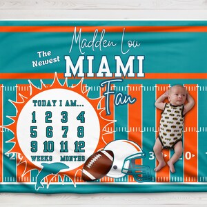 May include: A teal and orange baby blanket with a football theme. It features a football helmet, a football, a dolphin, and a sun with numbers 1-12 for weeks and months. The text "Madden Lou Miami Fan" is printed on the blanket.
