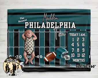 Puede incluir: Una manta de hitos con temática de fútbol en color verde azulado y negro, con un gráfico de campo, un balón, un casco y el texto The Newest Madden PHILADELPHIA Fan. Incluye un rastreador mensual de 1 a 12 meses y mide 127 cm por 152 cm.