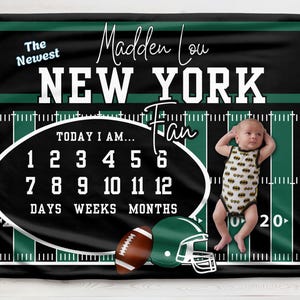 Puede incluir: Una manta de hito para bebé con temática de fútbol en negro y verde con el texto "The Newest NEW YORK Fan". Incluye un balón de fútbol, un casco y un espacio para rastrear la edad del bebé. Un bebé con un body de Batman está en la manta.