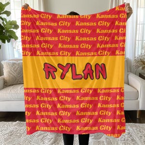 Pode incluir: Uma manta de lã vermelha e amarela com as palavras "Kansas City" repetidas em um padrão. O nome "RYLAN" está escrito em letras vermelhas grandes no centro da manta.