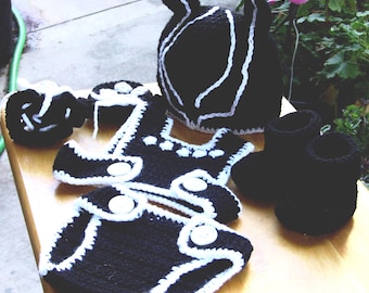 May include: A collection of handmade crochet baby items in black with white trim. The set includes a hat, booties, and diaper covers. The items are displayed on a wooden surface, showcasing the intricate crochet work and contrasting colors.