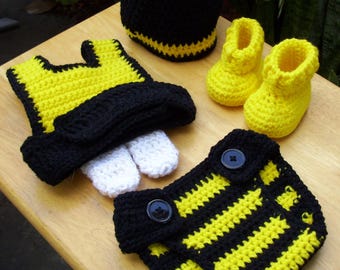 May include: A collection of handmade crochet baby items in black, yellow, and white. The set includes a beanie, booties, a diaper cover, a vest, and mittens. The items are arranged on a light brown wooden surface.