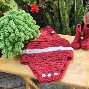 May include: A handmade crochet baby outfit set in red, white, and green. The set includes a romper with white accents, two red boots, and a green decorative item. The items are displayed on a wooden table.