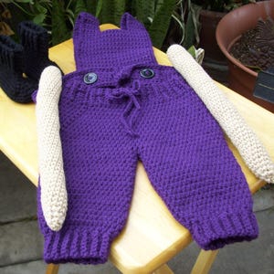 May include: A handmade purple crochet baby outfit with a hood and two large buttons. The outfit includes matching leg warmers and a pair of black crochet baby booties. The outfit is displayed on a wooden surface.