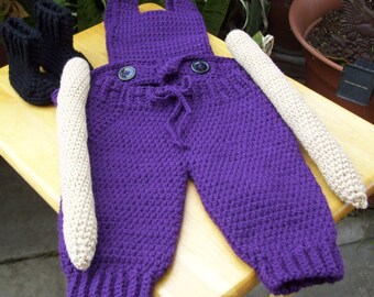 May include: A handmade purple crochet baby outfit with a hood and two large buttons. The outfit includes matching leg warmers and a pair of black crochet baby booties. The outfit is displayed on a wooden surface.