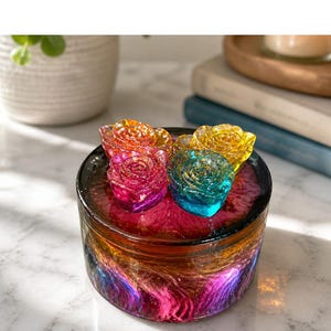Puede incluir: Un tarro decorativo redondo con un diseño arremolinado y colorido en tonos naranja, rosa, morado y azul. El tarro está rematado con cinco objetos en forma de rosa en varios colores, incluyendo naranja, rosa, amarillo y turquesa. El tarro está sobre una superficie blanca.